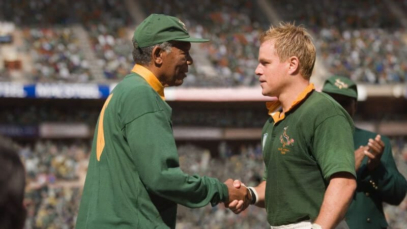 Dois homens se cumprimentando em um campo de rugby, os dois vestem uniformes verde e amarelo