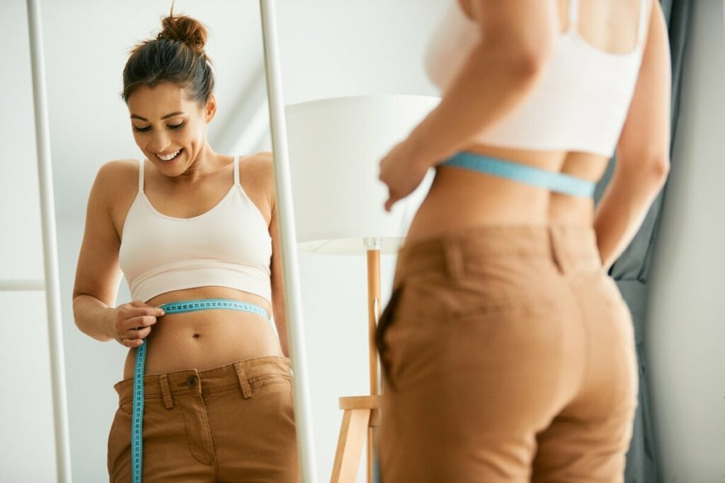 Mulher sorrindo em frente a um espelho medindo a cintura com fita