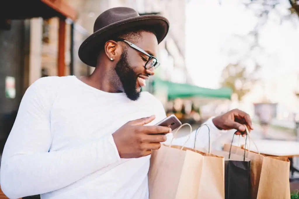 Homem mexendo no celular, sorrindo e segurando sacolas de compras