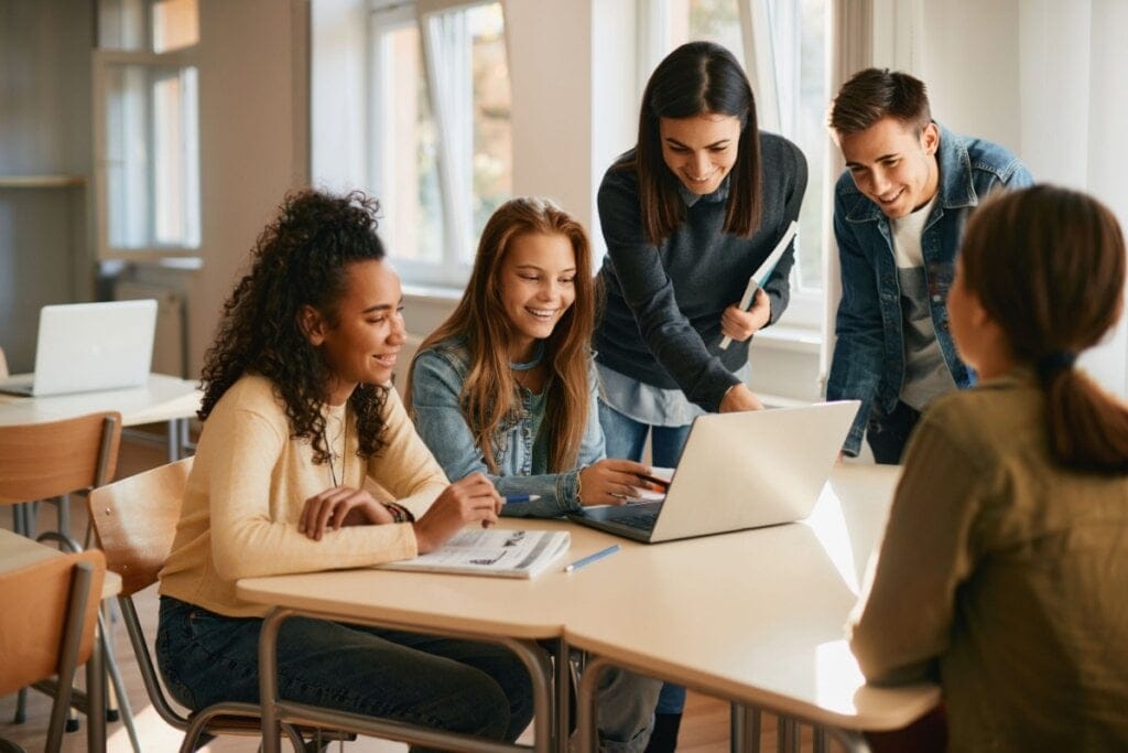 Estudantes sentado ao redor de uma mesa com professora em pé explicando algo em um notebook