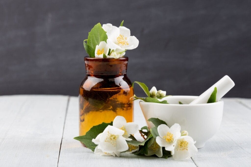 Vidro âmbar marrom com flores de jasmim dentro, em uma mesa branca, com recipiente com flores de jasmim