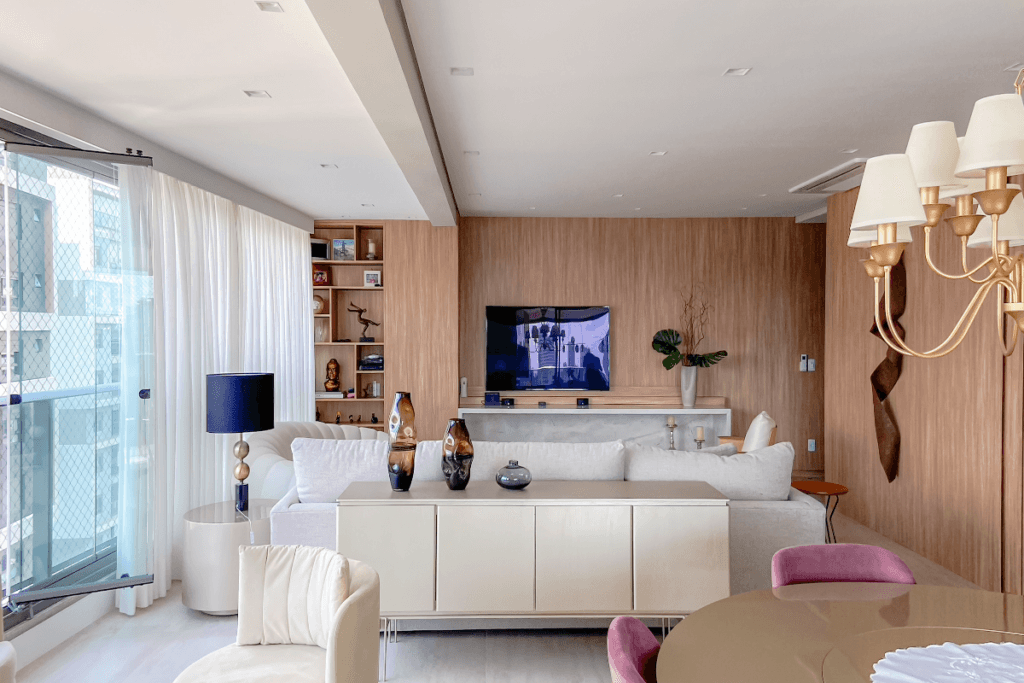 Sala de estar com móveis claros e buffet bege encostado nas costas do sofá com vasos em cima e itens de decoração, com uma parede de vidro e as demais em madeira, e a mesa da sala de jantar é bege escura com duas cadeiras estofadas em roxo e uma poltrona branca