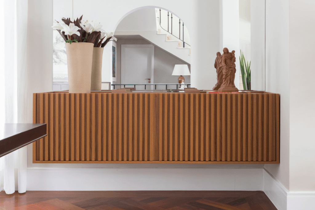 Parede branca com um buffet suspenso em madeira e com um espelho acima do móvel e com vaso e imagem de Nossa Senhora em cima do buffet e piso em madeira