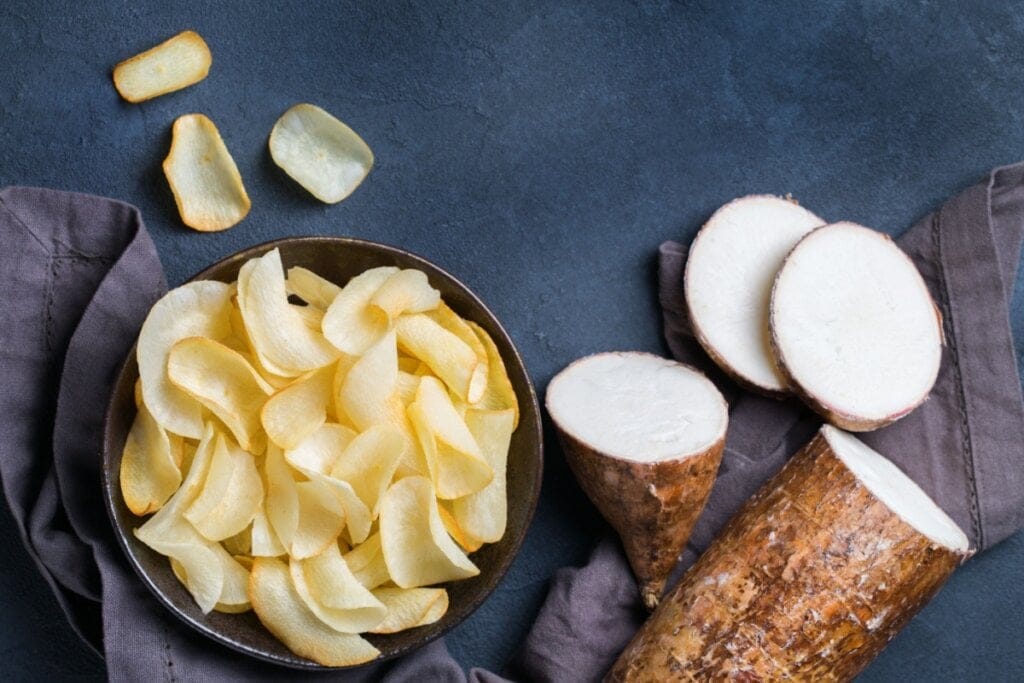 Tigela com chips de mandioca dourados ao lado de raízes cortadas