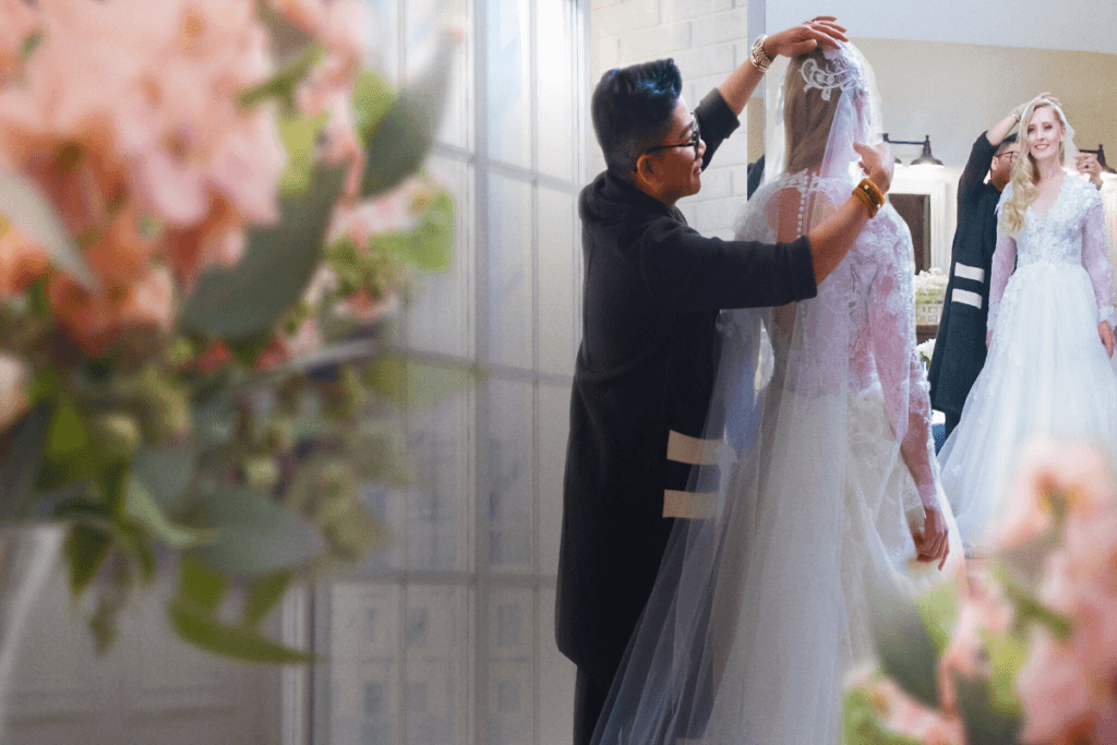 Mulher vestida de noiva em frente ao espelho, com homem com roupa social preta colocando o véu em sua cabeça