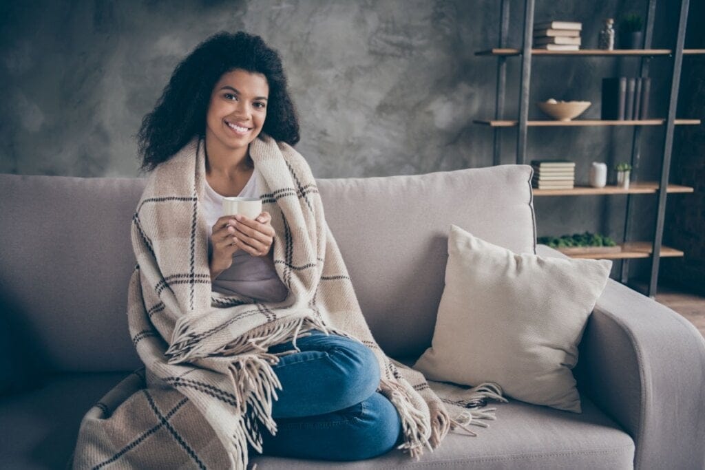 Mulher com cabelo cacheado solto vestindo blusa de frio e calça jeans sentada no sofá enrolada em manta e segurando uma caneca com chá