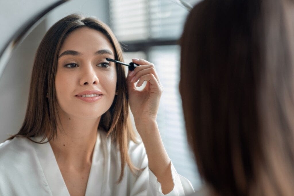 Mulher com o cabelo liso solto usando uma camisa branca em frente ao espelho passando uma máscara de cílios