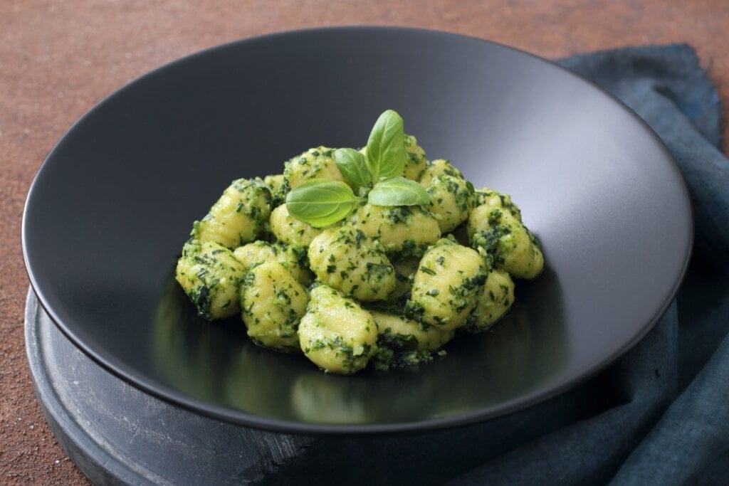 Nhoque ao molho pesto servido em prato fundo preto em cima de mesa de madeira