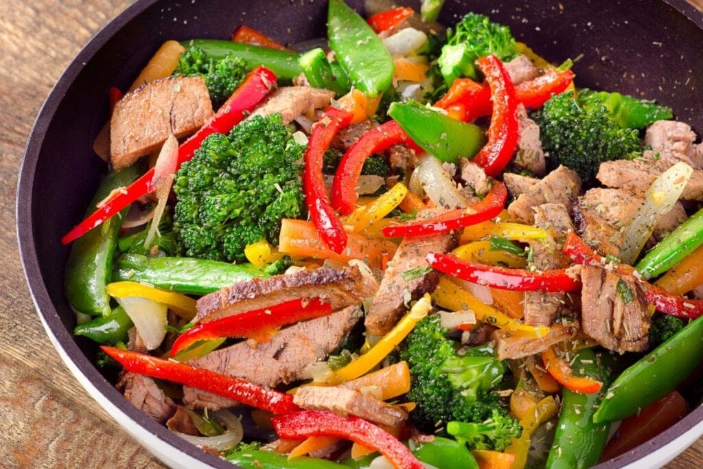 Tiras de carne com pimentão, ervilha e brócolis em uma panela preta 