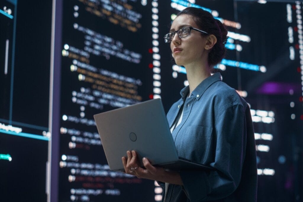 Mulher segurando um notebook e olhando para telas com linguagem de programação