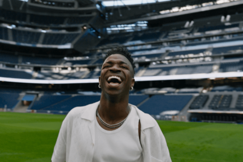 Homem negro com o cabelo curto, e usando regata e camisa branca sorrindo, no meio do gramado de estádio de futebol