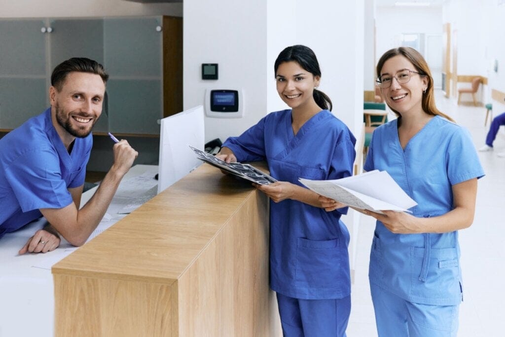 Homem com cabelo curto e barba, usando uma roupa de enfermagem azul, encostado em balcão de atendimento com outras duas mulheres, uma morena com cabelo castanho escuro e usando uniforme azul de enfermagem e outra mulher com cabelo claro, curto e usando óculos usando uniforme de enfarmagem azul