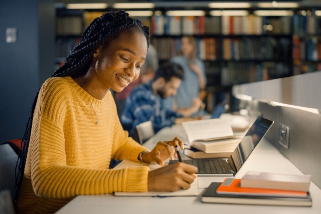 Estudante com o cabelo trançado, preso, usando um suéter amarelo escrevendo na biblioteca da universidade
