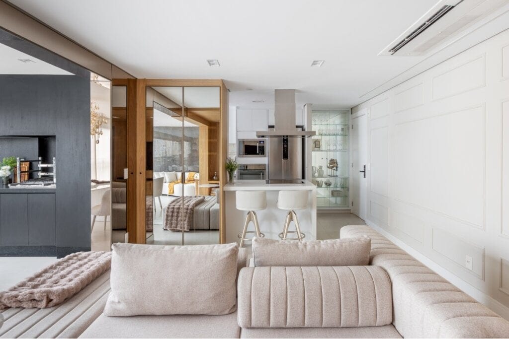 Sala de estar com um sofá bege na frente e, ao fundo, um móvel marrom com vidro e uma cozinha