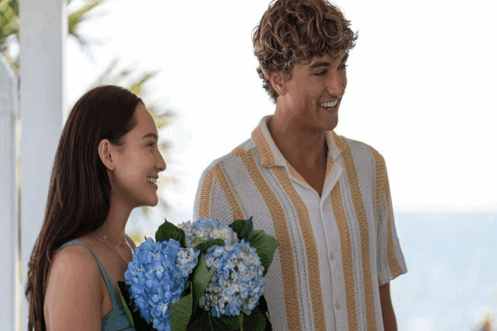 Imagem de uma jovem com o cabelo solto, usando um vestido de alcinhas verdes e com flores azuis nas mãos, ao lado um jovem com cabelo cacheado loiro, usando uma camisa listrada branca e laranja