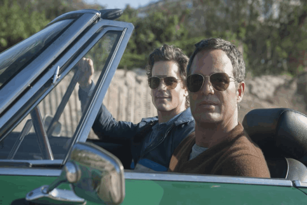 Imagem de dois homens dentro de carro com teto aberto,  um deles está usando óculos escuro e uma blusa marrom com camiseta branca e o outro está usando óculos escuro e uma blusa azul. 