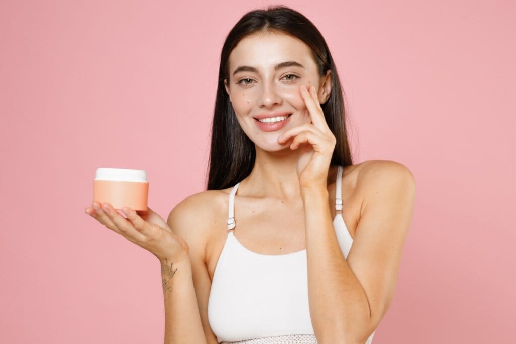 Mulher sorrindo, usando uma roupa branca em frente a um fundo rosa segurando um pote de creme em uma das mãos