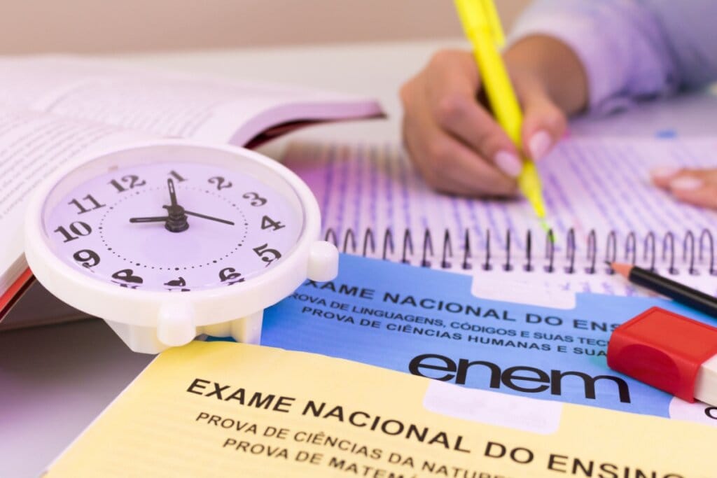 Relógio branco em cima de provas do Enem e uma mão com marca texto em cima de um caderno do lado
