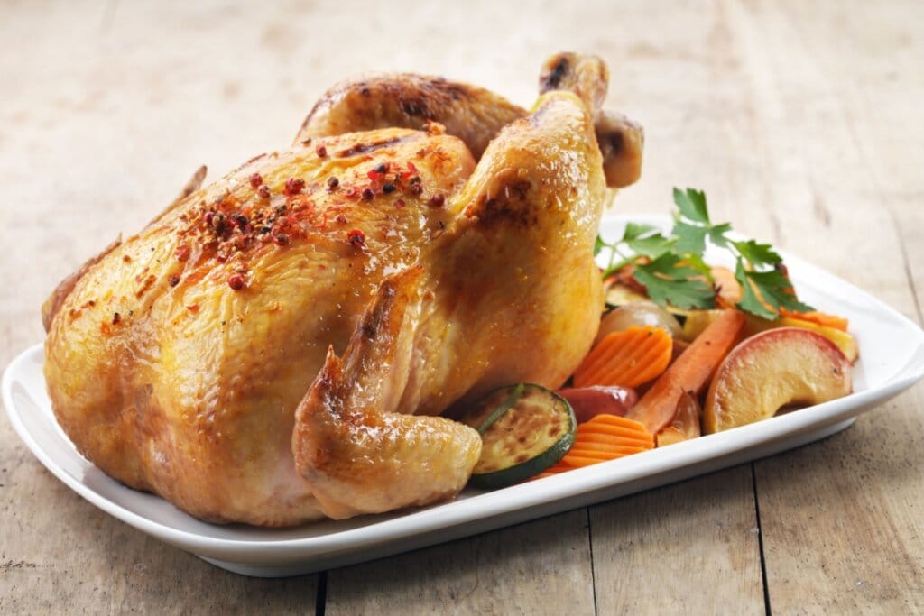 Travessa branca com frango assado e legumes, como cenoura, abobrinha e batata-doce