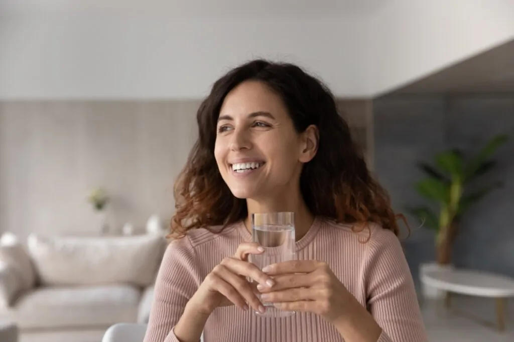 Mulher sorrindo, sentada e segurando um copo de vidro com água 