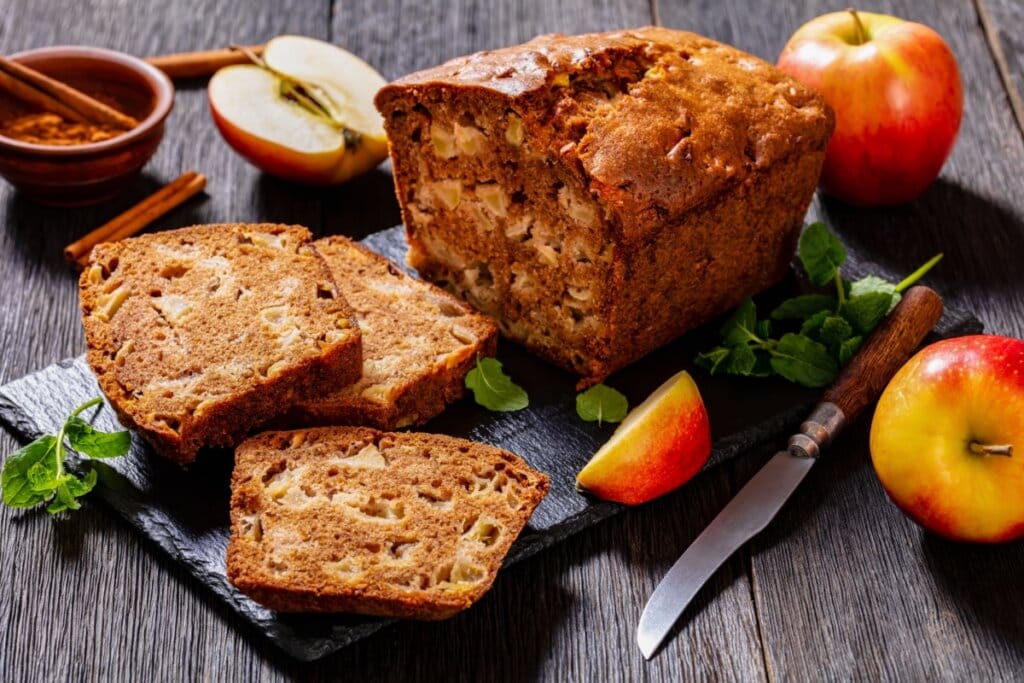 Pão doce de maçã fatiado, com pedaços visíveis da fruta e toque de canela, servido sobre tábua rústica com maçãs frescas ao redor