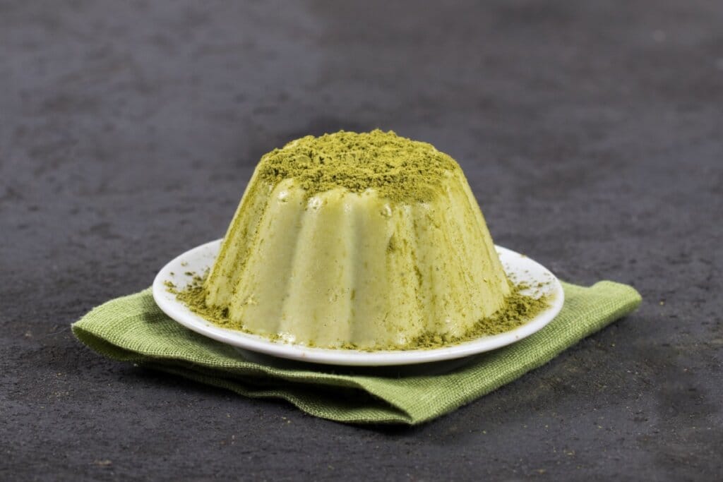 pudim verde com matcha em pó em cima em prato branco sobre pano verde