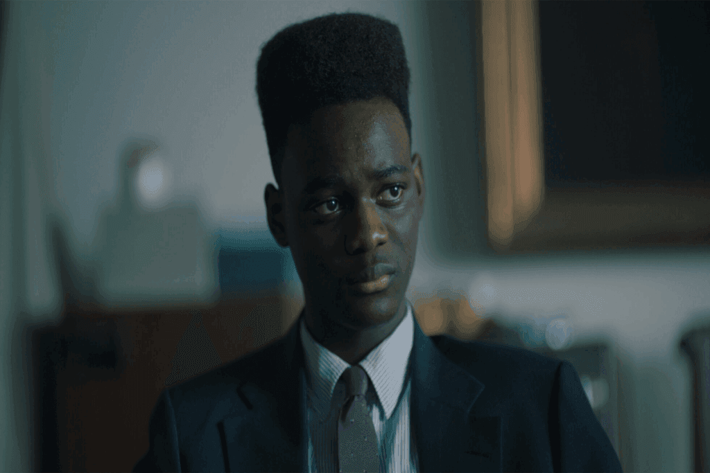 Jovem negro com o topete usando camisa social branca, gravata cinza e terno preto com uma expressão aflita