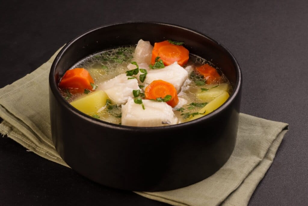 Sopa de peixe com cenoura e batata servida em tigela de cerâmica preta em cima de pano de prato cinza em cima de mesa de pedra