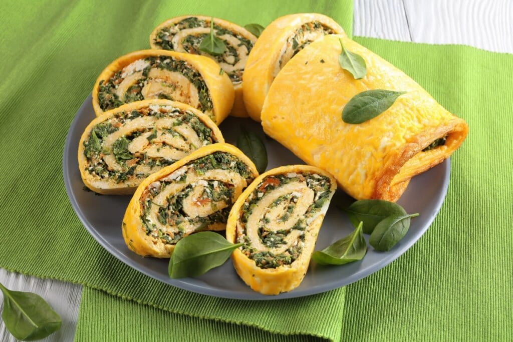 Rocambole de massa de ovos recheado com espinafre, cenoura ralada e queijo feta, servido em fatias sobre prato cinza com folhas frescas