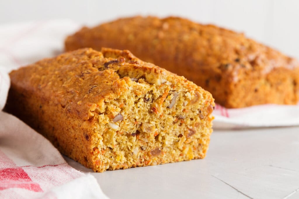 Bolo de cenoura e nozes cortada ao meio com bolo inteiro ao fundo em uma mesa branca 