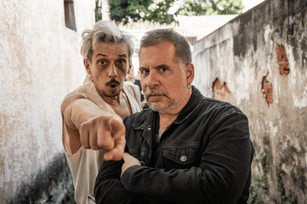Dois homens olhando para a câmera, um deles está usando jaquetas jeans preta e o outro está usando uma camiseta regata branca com o cabelo branco e bagunçado