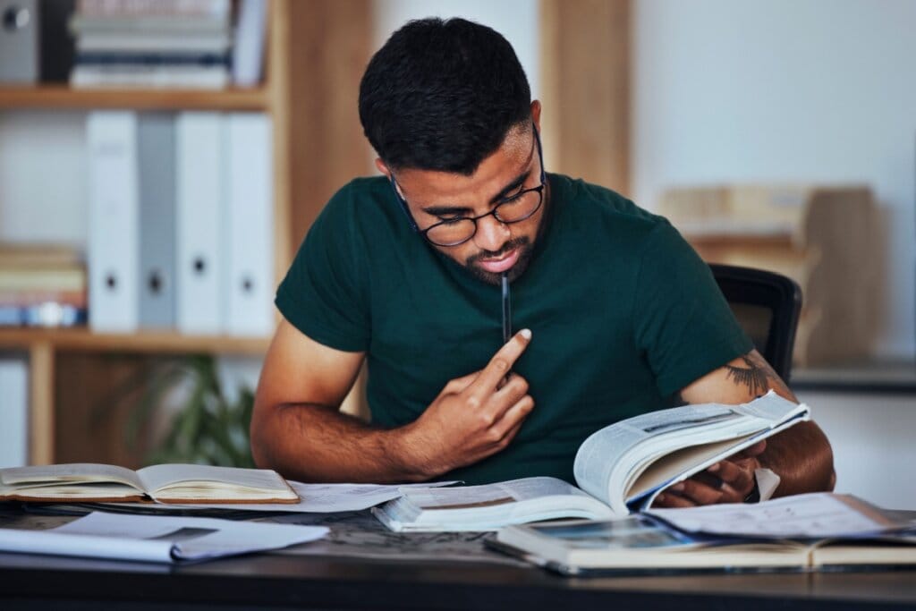 Jovem com cabelo curto, preto, usando camiseta verde-escura estudando livros