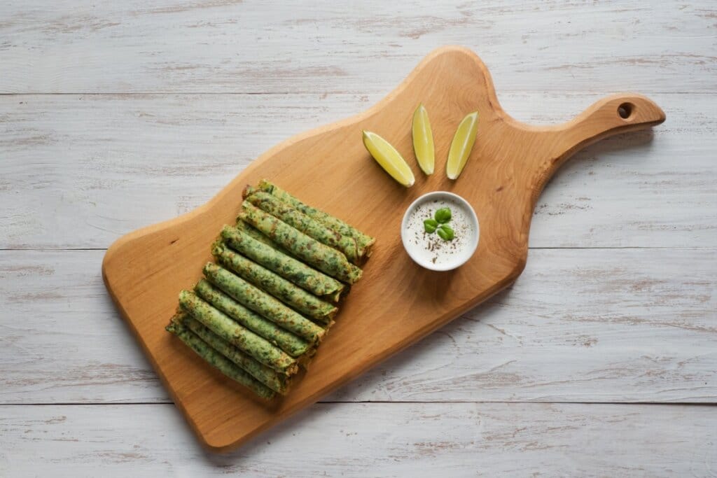 Panquecas de espinafres enroladas e empilhadas em cima de uma tábua de madeira. Ao lado, um recipiente com molho na cor branca e três fatias de limão 
