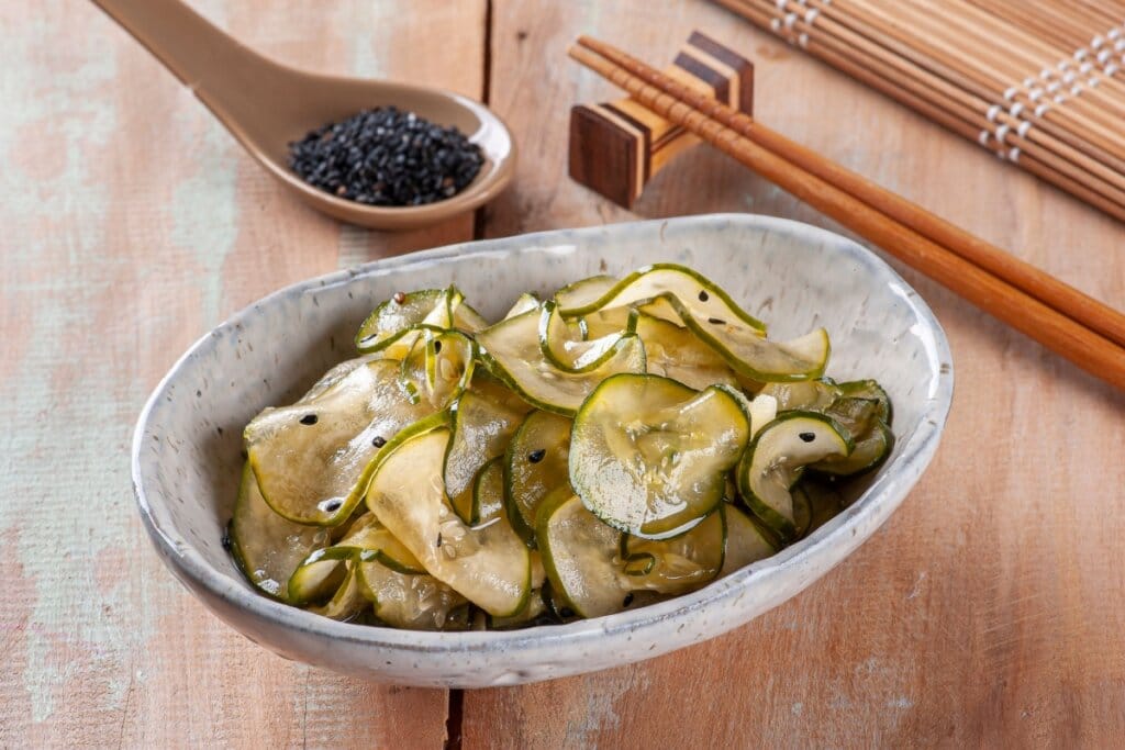 Uma tigela de sunomono com fatias finas de pepino temperadas e sementes de gergelim preto, acompanhada de hashis e colher de madeira