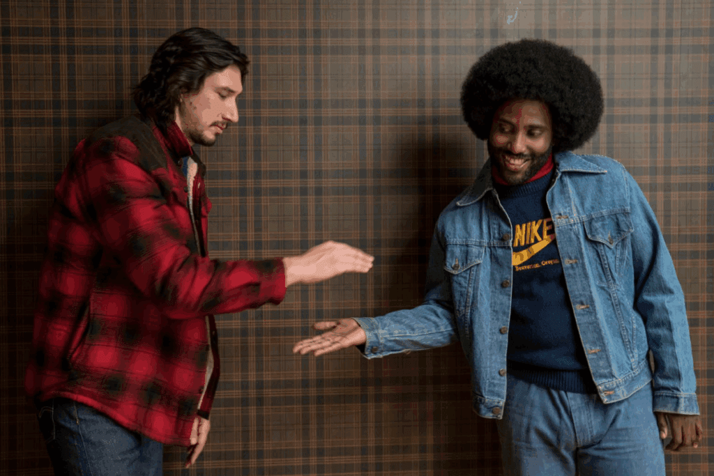 Dois homens, um de barba e casaco vermelho e outro de cabelo black e jaqueta jeans, fazendo um cumprimento amigável