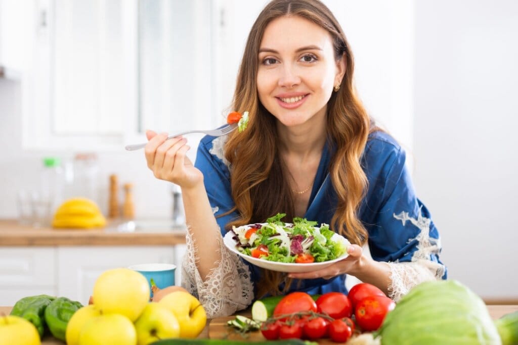 Jovem com o cabelo longo, castanho-claro, usando camisa azul com um prato de salada na mão e em cima da bancada há frutas e legumes
