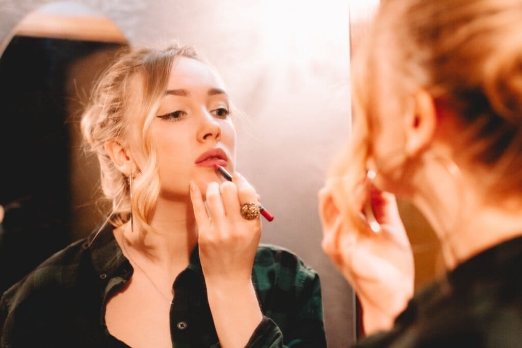 Mulher usando lápis labial para fazer contorno na boca em frente ao espelho, mulher tem o cabelo loiro e está usando uma camisa de botões preta