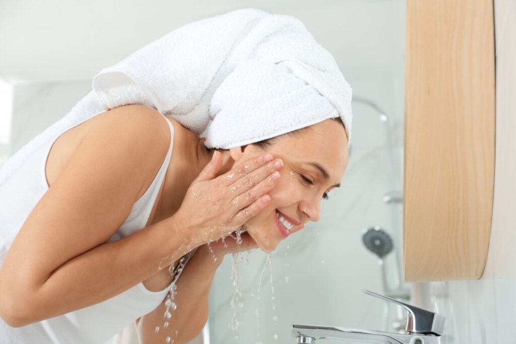 Mulher com toalha na cabeça e usando regata branca, lavando o rosto com água