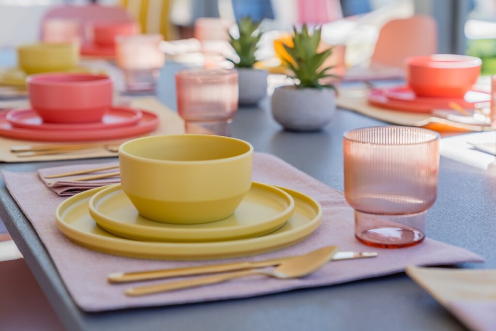 Mesa posta com pratos, talheres e copos coloridos, em tons de amarelo e rosa