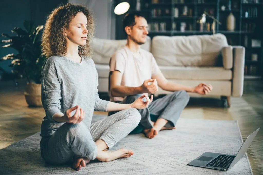 Homem e mulher meditando em posição de lótus na sala, com olhos fechados