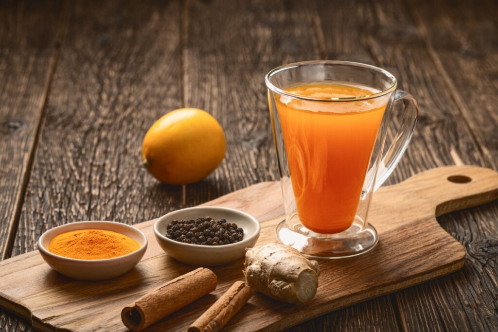 Copo de suco alaranjado em mesa rústica de madeira, acompanhado por manga, gengibre fresco, canela em pau, pimenta-do-reino em grãos e cúrcuma em pó