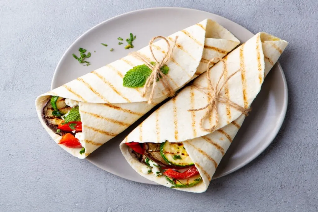 Dois wraps de legumes grelhados com queijo feta, amarrados com barbante e servidos em prato cinza