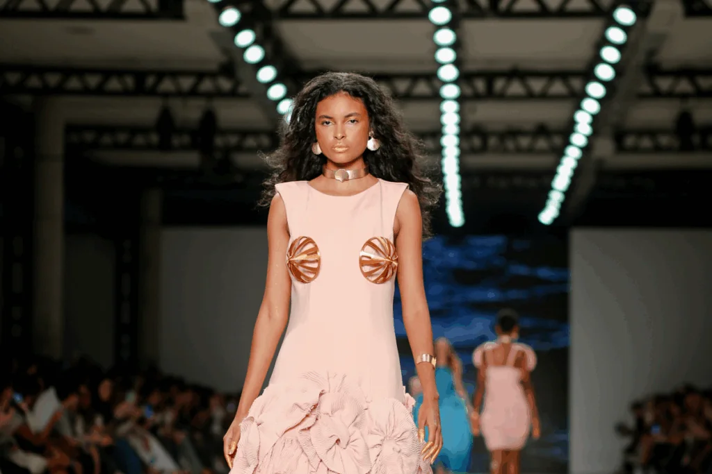 Mulher com o cabelo solto, e com movimento usando vestido rosa-claro com detalhes dourado em desfile de moda