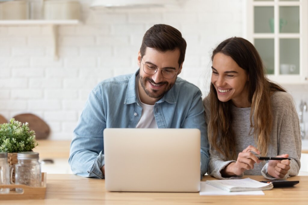 Casal sentado à mesa em frente ao notebook fazendo pesquisa. O homem tem o cabelo curto, usa óculos e barba, com uma camiseta branca e camisa jeans de botao e a mulher tem o cabelo longo, liso e solto. E está usando uma camisa de manga longa cinza
