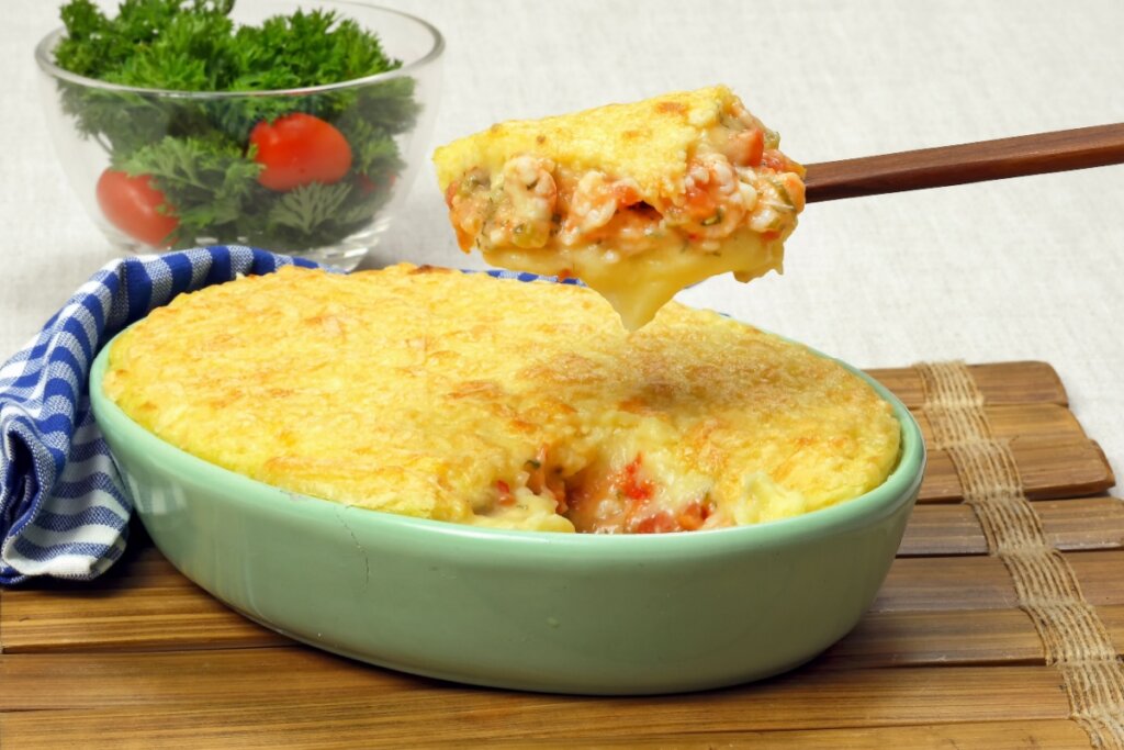 Escondidinho de camarão servido em refratário de cerâmica verde em cima de mesa de madeira com colher de madeira tirando um pedaço do escondidinho