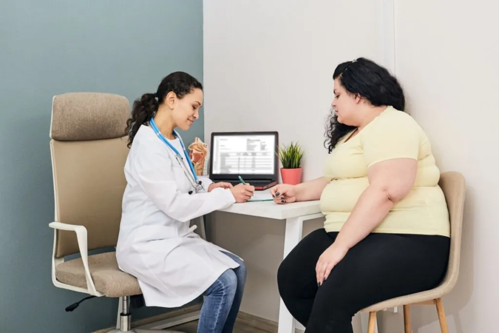 Médica de jaleco branco e paciente obesa de camiseta amarela sentadas em cadeiras em consultório