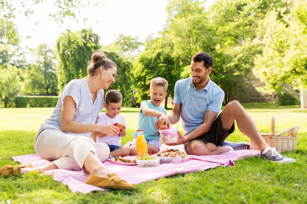 Mãe e pai com dois filhos pequenos fazendo piquenique em um parque, sentados sobre uma toalha cor-de-rosa, cercados por árvores e alimentos como suco e sanduíches