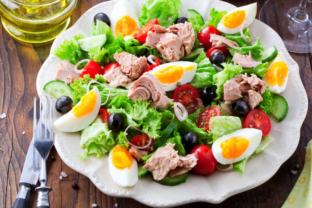Salada de atum com ovos e legumes servido em prato branco em cima de mesa de madeira