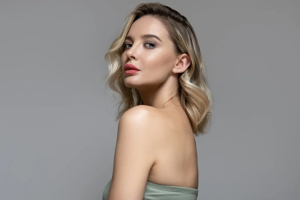 Mulher com cabelo loiro e raiz escura de lado, com top cinza e em fundo cinza