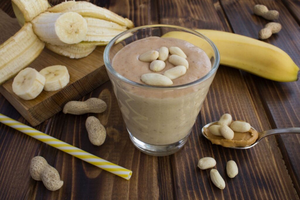 copo de vidro com vitamina marrom e amendoins em cima. em volta, amendoins, bananas fatiadas e colher com pasta de amendoim